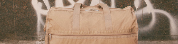 A large beige travel bag with handles and a front zipper pocket is placed against a wall with graffiti, in an urban outdoor setting.