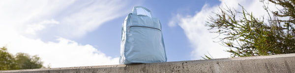 A small, vibrant pink crossbody bag is perched on a green metal fence, with blurred urban architecture in the background under natural daylight.