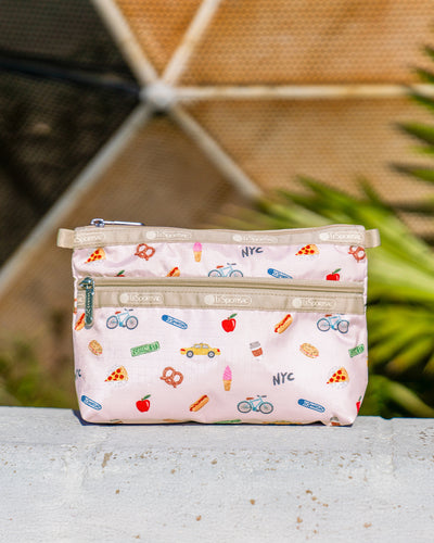 Cosmetic Clutch in NYC Icons Pink featuring playful illustrations of pizza, pretzels, taxi cabs, bicycles, apples, and “NYC” lettering on a pale pink background with Latte trim and zip closures.