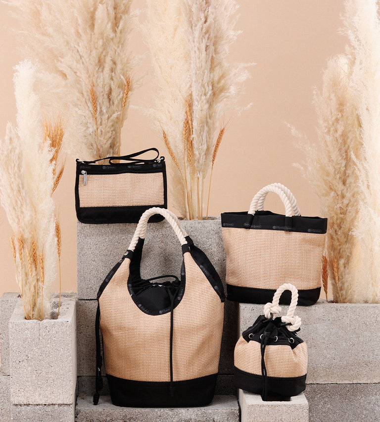 Four Raffia-textured LeSportsac bags with black nylon accents are artfully arranged on cement blocks, surrounded by tall pampas grass against a warm beige background. The display includes a variety of styles, featuring rope handles and drawstring details, evoking a relaxed, natural aesthetic.