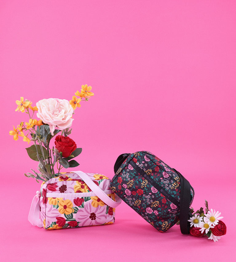 Two floral-patterned LeSportsac crossbody bags are displayed against a bright pink background, one in pastel tones with large flowers and the other in darker tones with intricate floral details. Flowers are arranged inside and around the bags, enhancing the spring-inspired aesthetic.
